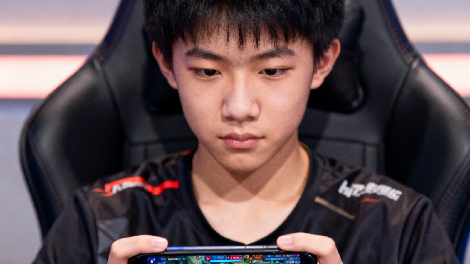 一位约17岁的青训选手坐在专业电竞椅上，双手熟练地操作手机屏幕进行训练赛，表情专注而镇定，屏幕显示着复杂的游戏界面，背景虚化处理凸显人物主体，展现新一代电竞人才的培养场景。